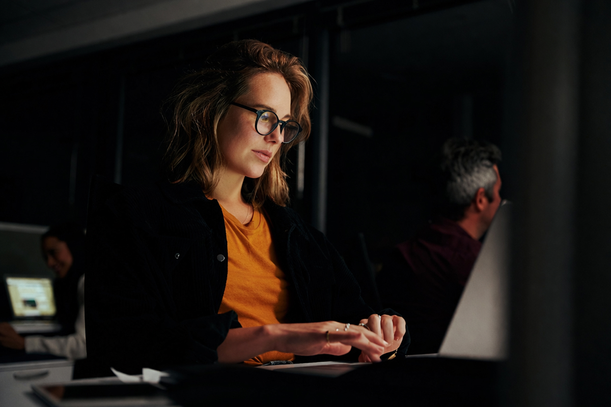 A woman wearing glasses typing on a laptop in a dark setting.